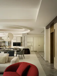 Modern open-plan living room, dining area and kitchen featuring a curved white marble island, round black marble dining table, sculptural white pendant light, neutral sofas, and a vibrant red curved armchair in the foreground.