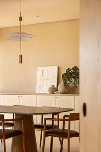 Modern minimalist dining room featuring a round oak pedestal table, wooden chairs with brown leather seats, a sculptural white textured artwork, ceramic vases with eucalyptus, and a two-tier white pendant light against a textured beige wall.