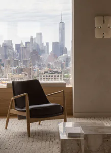 Modern oak and black leather armchair placed on a textured rug in front of a large window overlooking the New York City skyline with One World Trade Center prominent, next to a marble coffee table and a modern wall sconce.