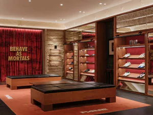 Luxury shoe boutique interior with warm walnut wood display cabinets, black leather benches, red velvet curtain with gold 'BEHAVE AS MORTALS' lettering, and orange carpet with 'PANE' branding.