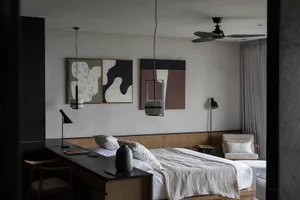 Modern minimalist bedroom interior with a wooden platform bed, dark wood desk and chair, abstract green and brown wall art, black pendant lights, and a black ceiling fan.