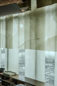 Interior view of a minimalist cafe featuring hanging translucent washi paper noren curtains with organic water-stain patterns, wooden counter with stacked ceramic dishes and trays, and a view of an outdoor bamboo patio with wooden furniture.