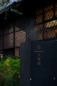 A modern black metal sign pillar featuring a bronze Chinese knot logo, the text DAQIANLOFT, and large Chinese characters 大錢閣, set in front of a dark building with decorative patterned metal screens and lush green bushes.