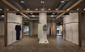 寬敞的極簡快閃店內部，展現裸露木結構樑柱、混凝土牆面、淺色木地板，以及懸掛在黑色衣架與中央展示桿上的設計師服裝。