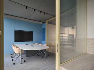 Modern conference room with bright blue textured wall, large black monitor, white curved meeting table, beige office chairs on casters, and a large glass partition wall with yellow frame.