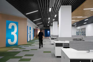 Modern office interior hallway with large blue room number 3 sign, green and grey checkerboard carpet tiles, white workstations with grey mesh chairs, linear ceiling lights, and a person walking toward the background.