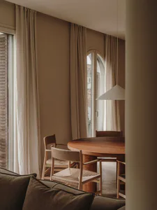 A warm minimalist dining room featuring a round walnut wood table, four light wood cord chairs, neutral beige curtains, and a white pendant lamp.