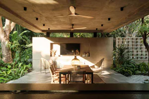 Modern tropical outdoor dining area under a raw concrete canopy with wooden dining table, woven chairs, ceiling fans, and abundant tropical plants.