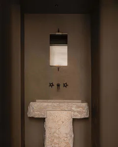 A minimalist bathroom featuring a thick travertine stone console sink with a matching towel holder, dark bronze cross-handle faucets, and a vertical rectangular mirror mounted on a textured taupe wall.