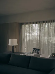 Modern minimalist living room interior featuring a dark teal sofa in the foreground, a wooden table with an open book and table lamp, a woven black leather chair, floor-to-ceiling sheer curtains, and warm wood paneling under soft natural light.