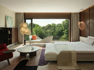 Modern luxury bedroom interior with large bed, red armchair, sculptural floor lamp, white chaise lounge, and expansive window overlooking a dense green forest.