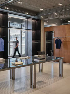 Modern industrial-style clothing boutique interior featuring a large glass-top display table with stainless steel frame, dark metal wall panels, warm wood accents, and concrete ceiling with exposed ductwork.