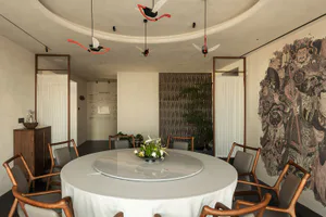 Modern Chinese-style dining room with a large round marble lazy Susan table, wooden dining chairs with grey upholstery, dramatic Tang dynasty mural on the right wall, and artistic red and white ceiling fans hanging from a curved ceiling.