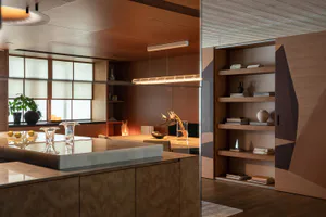 Modern dining room interior with a large stone-topped kitchen island, warm wood cabinetry, built-in geometric bookshelves, and a long cylindrical pendant light over the dining table.
