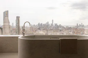 A modern concrete freestanding bathtub with a curved metallic faucet positioned in front of a large window overlooking the New York City skyline, including the Empire State Building and One World Trade Center.