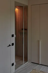 Modern minimalist walk-in steam shower with travertine stone walls, warm ambient lighting from above, glass door, built-in shelf with toiletries, and adjacent beige cabinetry in a neutral-toned bathroom.
