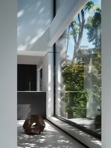 A bright, minimalist modern living room interior with white walls, large floor-to-ceiling windows overlooking green trees, a dark wood counter in the background, and a distinctive dark wooden sculptural stool on the floor in the foreground.