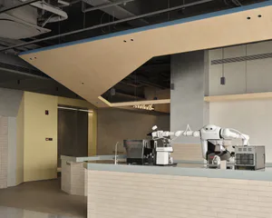 Modern coffee shop interior with a white robotic barista arm operating an espresso machine behind a white brick counter, under a large wooden architectural canopy with exposed ceiling ducts.