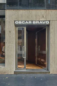 Modern minimalist storefront entrance featuring raw concrete facade, large stainless steel framed glass doors, and an illuminated Oscar Bravo sign.