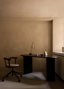 A minimalist home office corner featuring a dark walnut curved desk, matching swivel armchair with wheels, a sculptural black desk lamp, and various small decorative objects against warm beige plastered walls.