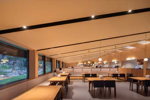 Modern minimalist restaurant interior featuring extensive light wood finishes, built-in banquette seating along large windows, wooden dining tables with black chairs, and warm ambient lighting.
