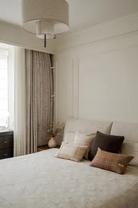 Modern neutral bedroom interior with cream textured bedspread, multiple pillows in beige, brown and plaid patterns, large fabric pendant light, and soft natural window light.