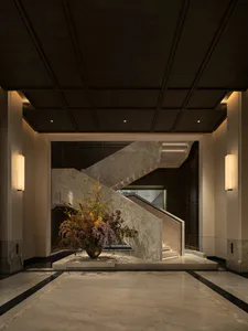 View of a luxurious hotel lobby featuring a dramatic white marble grand staircase with integrated lighting, a large bronze vase with an elaborate dried floral arrangement in autumn tones, dark coffered ceiling, and polished marble floor with black inlays.