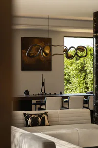 Modern dining area featuring an organic-shaped brass and dark metal chandelier, dark wood table, cream upholstered chairs, a white sofa with abstract black and beige pillow in the foreground, and a large abstract painting on the wall.