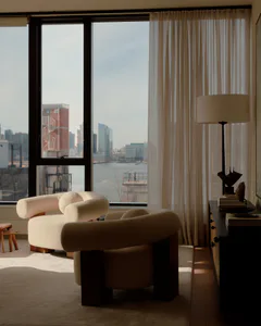 Modern living room interior featuring two cream boucle armchairs with dark wood bases, a black console table with a tall floor lamp, sheer curtains, and a large window revealing a panoramic view of the New York City waterfront with brick buildings and the river.
