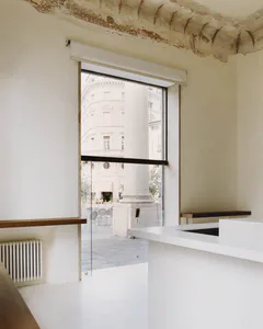 Modern minimalist white kitchen interior with wooden shelves, white marble counter, and large glass opening revealing an ancient stone column and classical building outside.