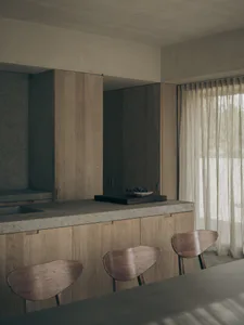 Modern minimalist kitchen featuring a concrete countertop island, light oak cabinetry, wooden butterfly chairs, and a dark concrete accent wall with a bowl of fruit on a wooden cutting board.