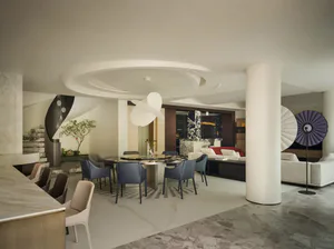 Modern luxury open plan interior featuring a round marble dining table with blue velvet chairs, sculptural white chandelier, white curved ceiling details, and living area with white sofas in the background.