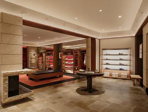 Interior view of a high-end shoe boutique with a large travertine fireplace on the left, built-in wooden display shelves with red interiors holding various shoes, a central round dark wood table on a circular rug, and additional floating shelves on the right wall displaying more footwear.