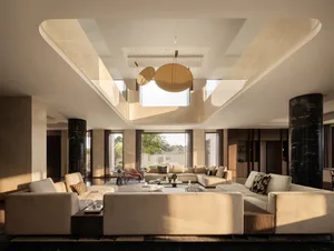 Spacious modern luxury living room featuring a double-height ceiling with skylights, large beige sectional sofas, black marble cylindrical columns, and a sculptural pendant light fixture.