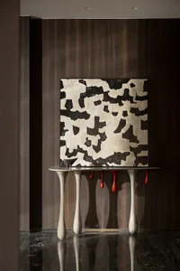 Modern interior scene featuring a sculptural off-white console table with four curved legs, a glossy black marble floor, dark wood paneling, and a large textured black and white abstract painting hanging above it. Red drip-like sculptures hang from the table surface.