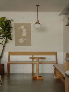 Minimalist café interior with a light oak wooden bench and table set against a white tiled wall, a hanging fabric banner with coffee equipment illustrations, a large fiddle leaf fig plant, and a white glass pendant light.