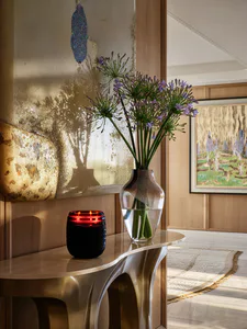 Curved brushed-brass console table displaying a glass vase of purple agapanthus flowers, a glowing red candle holder, and an abstract textured painting in a sunlit entryway.