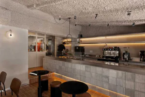 Modern minimalist cafe interior with a long grey tiled counter, black round tables, brown leather stools, and a striking textured concrete ceiling with pendant lights and spotlights. A barista is working behind the counter with professional espresso machines visible.