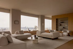Modern minimalist living room in a high-rise apartment with beige modular sofas, a round travertine coffee table, wooden built-in shelving, and floor-to-ceiling windows overlooking a city skyline and river.