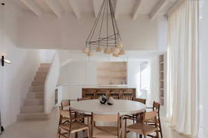 Modern minimalist dining room featuring a large round travertine table surrounded by six wooden woven chairs, with a sculptural black pendant light above, white walls, a curved staircase on the left, and an open kitchen with wooden bar stools in the background.
