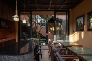 Semi-outdoor dining area featuring a curved black terrazzo bar counter, wooden dining tables with green tiles, terracotta wall tiles, and a lush courtyard with trees visible through large glass doors.