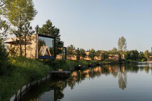 Modern wooden cabins with large glass windows and black frames elevated over a calm lake surrounded by pine trees, with clear reflections in the water.