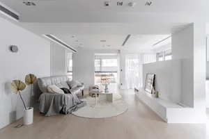 Modern minimalist living room with white walls, light wood flooring, grey leather sofa, round white rug, and large dried palm leaves in a white vase.