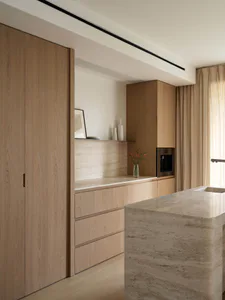 Modern minimalist kitchen featuring light oak wood cabinetry, a large travertine kitchen island, built-in appliances, and soft natural lighting.