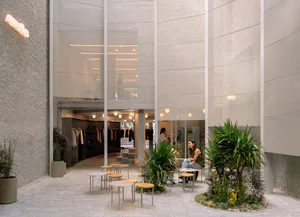 Modern retail atrium featuring textured concrete walls, large glass panels, indoor plants, and circular tables with people relaxing in a bright, open space.