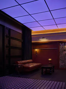 Interior of a stylish speakeasy lounge featuring a brown leather daybed, dark wood paneling, purple grid ceiling with LED lighting, and a tropical leaf wallpaper panel.