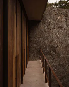 Narrow outdoor corridor featuring vertical teak wood cladding on the left and a rough stone wall on the right, with a metal handrail overlooking the edge.