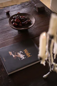 A dark wooden table featuring a shallow wooden bowl filled with dried persimmons, a black book titled Wabi Sabi, and a lit white candle with melted wax in the foreground.