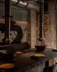Industrial-style hotpot restaurant interior featuring dark concrete walls, large stainless steel exhaust ducts, and a black stone counter set with hotpot pots and wooden plates.