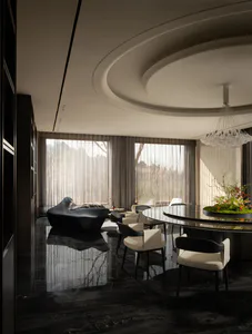 Modern luxury dining and living area featuring a large round black marble table, cream and black dining chairs, a sculptural black chaise lounge, dramatic curved ceiling with a large crystal chandelier, and floor-to-ceiling windows overlooking a forest view.